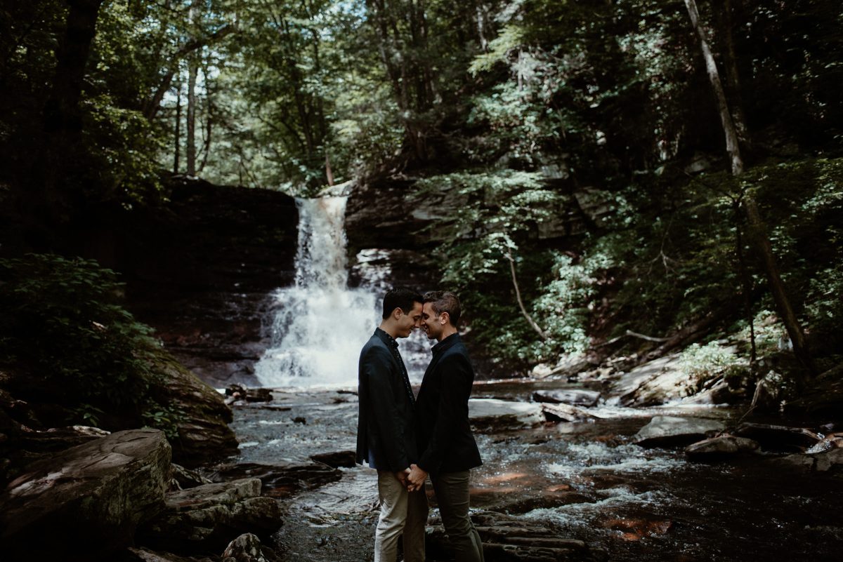 Two partners standing together by a waterfall during their LGBTQIA+ engagement session in the Pocono Mountains.