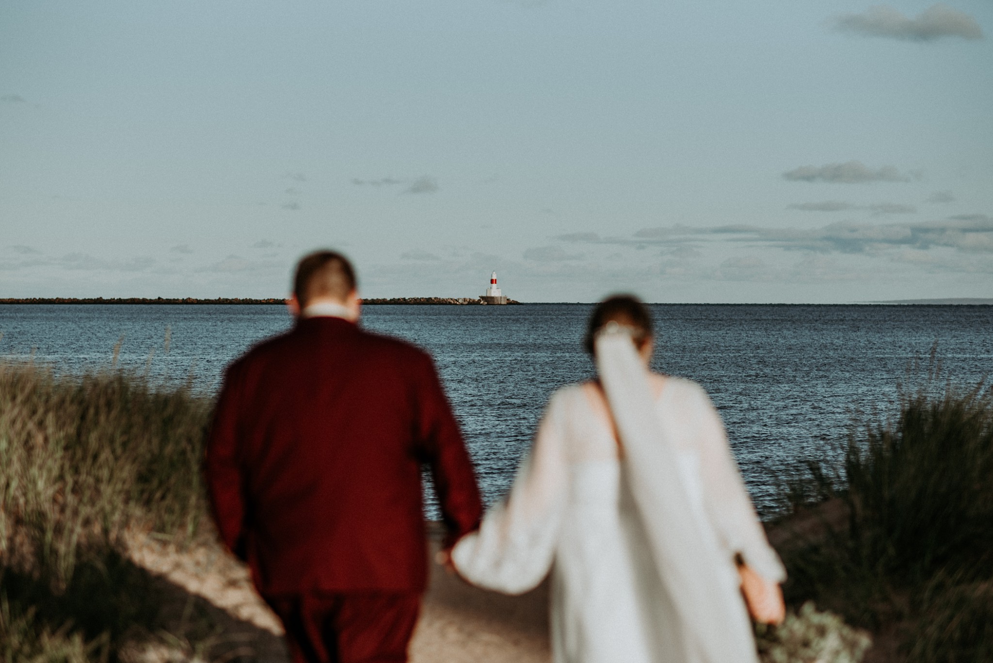 A bride and groom walking towards a lighthouse in Marquette.