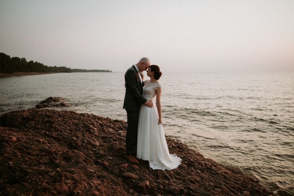 Bride and groom standing close together on rocky Lake Superior shoreline during Michigan elopement, groom touching bride’s face lovingly