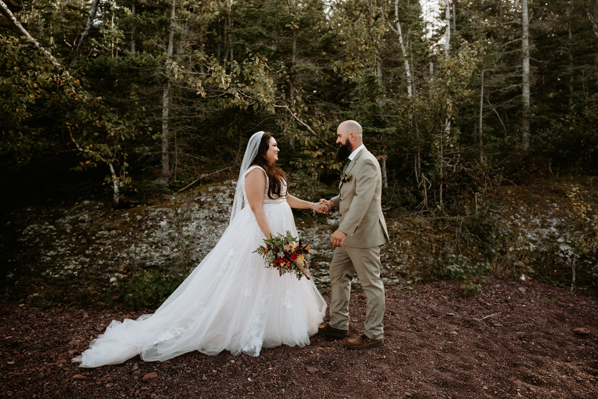 Bride and groom holding hands in forest clearing during Michigan elopement, bride wearing flowing white gown and holding summer bouquet