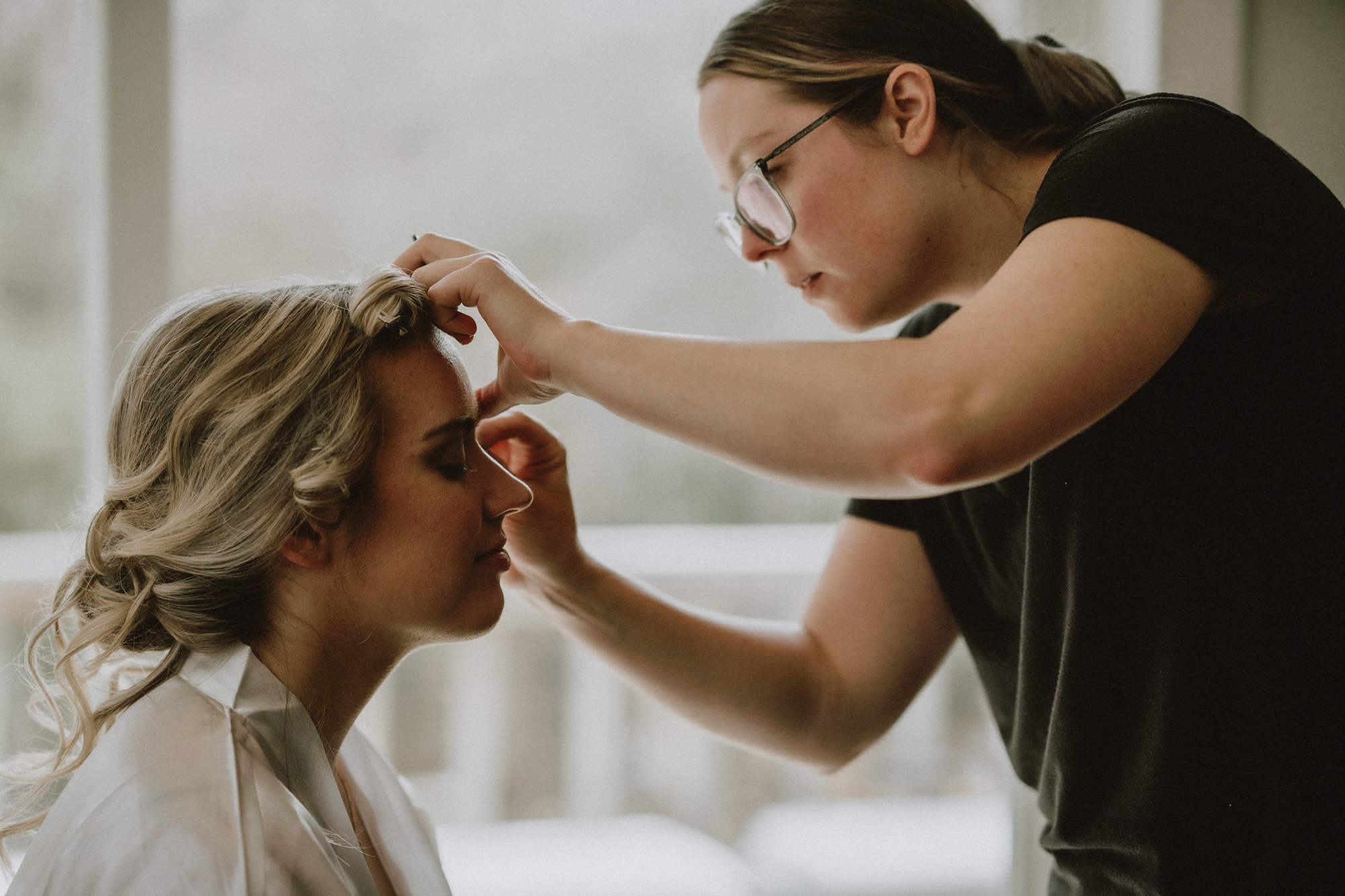 Bride having makeup touched up before Michigan elopement ceremony, natural light streaming through window